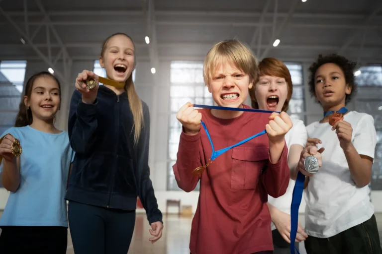Fünf Kinder stehen beieinander, halten Medaillen in den Händen und strahlen über das ganze Gesicht. Zwei Kinder links halten Goldmedaillen, zwei rechts Silber- und Bronzemedaillen. Das Kind in der Mitte hält eine Goldmedaille an einem blauen Band, lächelt breit, wirkt aber angespannt. Sie freuen sich riesig über ihren Sieg.