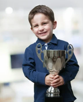 chłopiec uśmiecha się i trzyma trofeum. Ma na sobie granatową koszulkę polo. Trofeum jest srebrne z czarną podstawą.