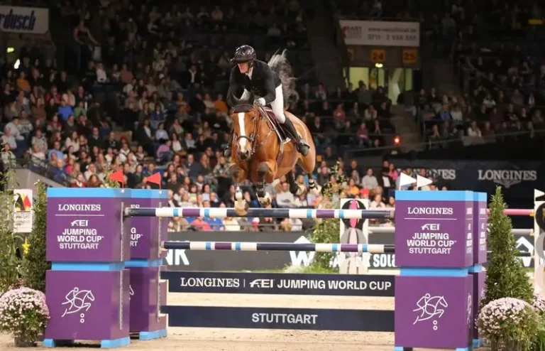 Ein Reiter mit schwarzem Helm und Springjacke überwindet auf einem braunen Pferd mit weißer Blesse ein hohes Hindernis beim Springreiten im Rahmen des Longines FEI Springweltcups in Stuttgart. Das Hindernis trägt das Longines-Logo und die Aufschrift „FEI Springweltcup Stuttgart“. Im Hintergrund ist die vollbesetzte Tribüne der Hanns-Martin-Schleyer-Halle mit zahlreichen Zuschauern zu sehen.