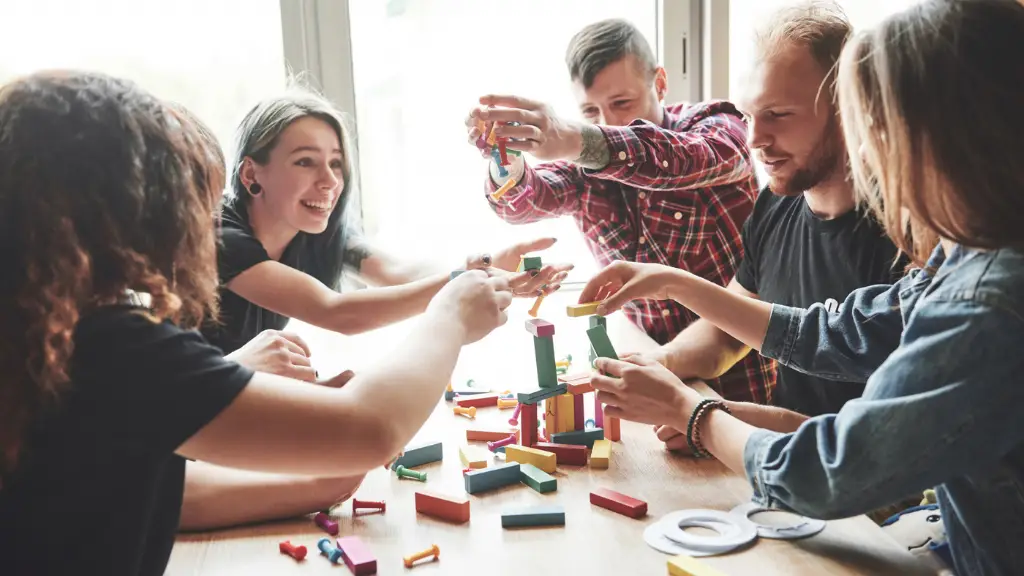 Eine Gruppe von Kollegen sitzt bei einem Teambuilding-Event um einen Tisch und spielt Jenga. Ein wackeliger Turm aus bunten Holzklötzen steht auf dem Tisch, und die Teilnehmer ziehen und stapeln die Klötze vorsichtig, lachen und konzentrieren sich auf das Spiel. Im Hintergrund ist ein Fenster mit Tageslicht zu sehen.
