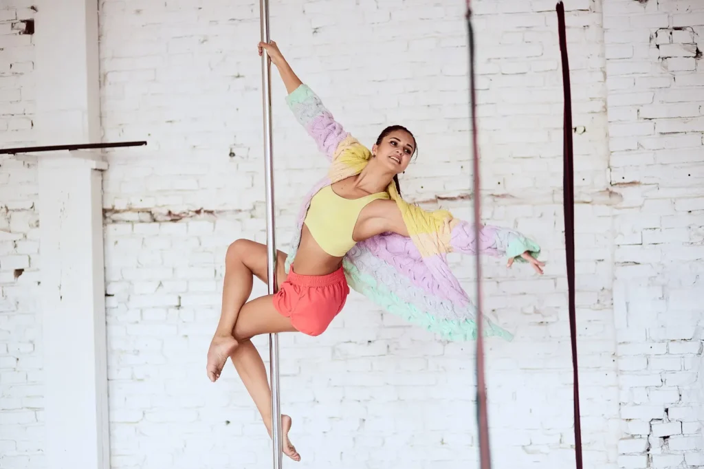 Eine Person übt Pole Dance in einem hellen Loft-Interieur. Sie trägt ein Sporttop, Shorts und einen farbenfrohen Pullover und vollführt eine beeindruckende Pose an der Stange.