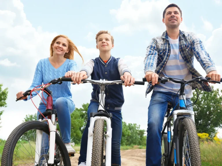 Eine Familie – eine Frau, ein Junge und ein Mann – fährt an einem sonnigen Tag nebeneinander mit Mountainbikes auf einem Waldweg. Alle drei blicken in die Kamera und sehen sehr glücklich aus.