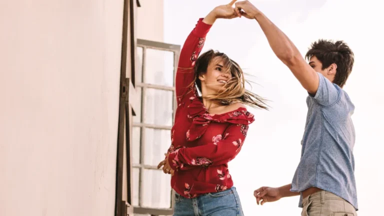 Eine Frau in einer roten Bluse mit Blumenmuster und Jeansshorts hebt lächelnd die Hand, ihr Haar weht im Wind, während sie tanzt. Ein Mann in einem blauen T-Shirt und beigen Hosen hat ebenfalls die Hand erhoben und führt die Frau vermutlich beim Tanz. Der helle Hintergrund lässt vermuten, dass sie im Freien oder an einem gut beleuchteten Ort tanzen. Die Atmosphäre ist fröhlich und lebhaft.