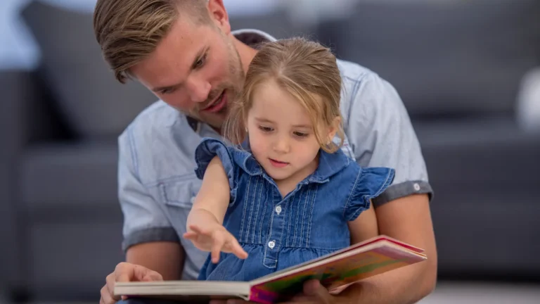 Ein Mann liest einem kleinen Mädchen ein Bilderbuch vor. Er hat kurzes braunes Haar und einen kleinen Bart. Er trägt ein hellblaues Hemd. Das Mädchen hat ebenfalls hellbraunes Haar und trägt ein Jeanskleid. Sie sitzt auf seinem Schoß und schaut in das Buch. Er zeigt auf etwas auf der Seite.