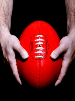 Nahaufnahme von Händen, die einen roten Australian-Rules-Football vor schwarzem Hintergrund halten.