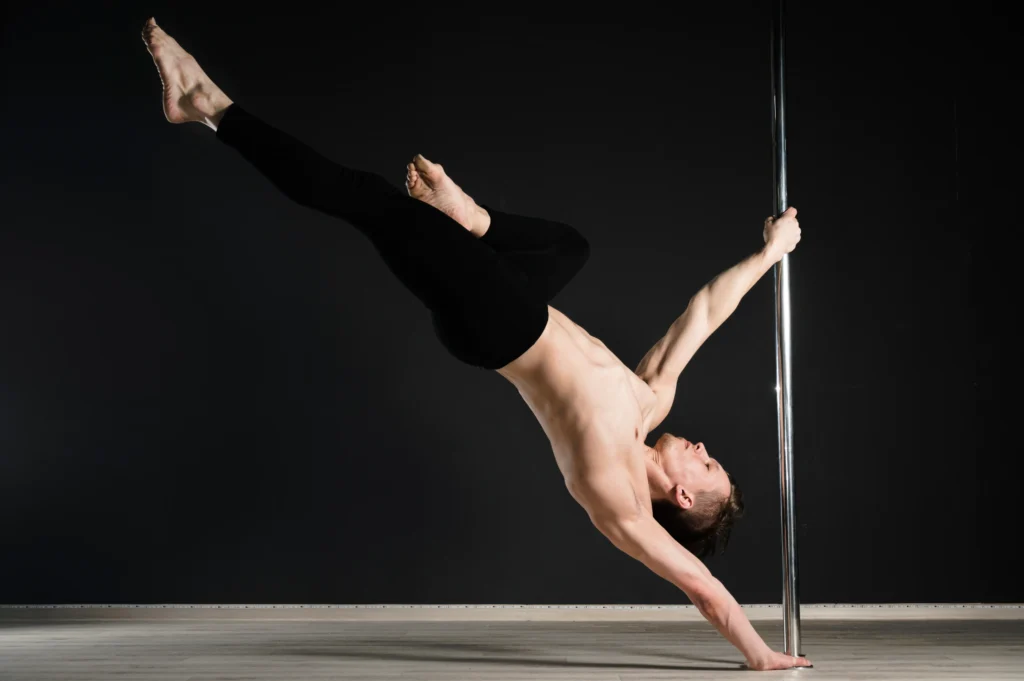 Ein männlicher Pole-Dancer vollführt einen Attitude Spin an einer vertikalen Metallstange. Er trägt schwarze Leggings und sein Oberkörper ist frei. Sein Körper ist in einer dynamischen Pose gebeugt, ein Arm hält die Stange, der andere ist nach unten zum Boden ausgestreckt, die Beine sind in der Luft. Der Hintergrund ist dunkel, der Boden hell.