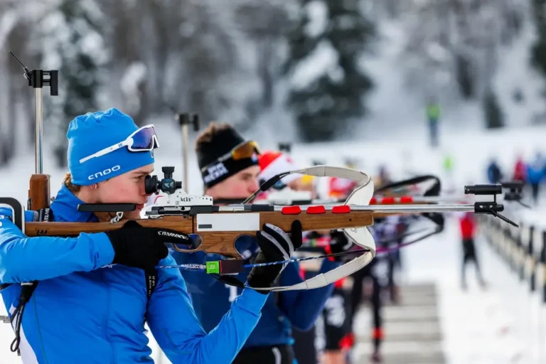 Biathlon-Wettkämpfer schießen während eines Winterwettbewerbs mit Gewehren auf Zielscheiben; im Hintergrund sind eine schneebedeckte Bahn und andere Athleten zu sehen.