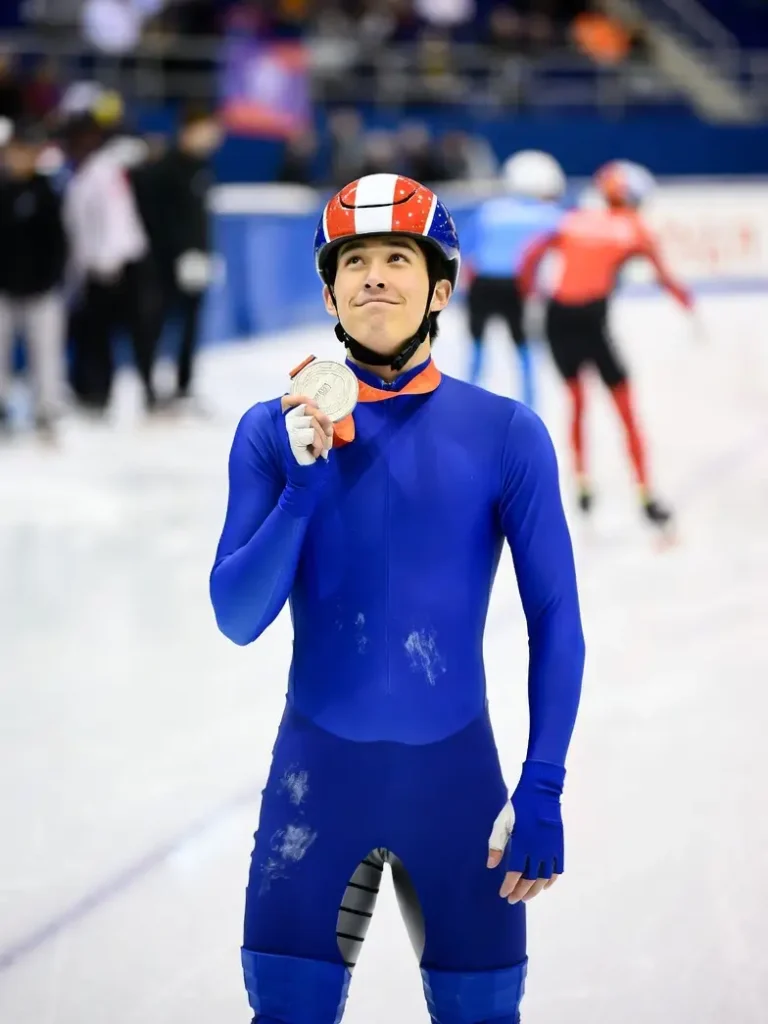 Ein Shorttrack-Eiskunstläufer nach seinem Rennen – ein junger Läufer in blau-rot-weißem Anzug und Helm, der eine olympische Silbermedaille in der Hand hält. Er lächelt leicht, den Kopf erhoben, und steht auf der Eisfläche, umgeben von den Tribünen der Olympiahalle.