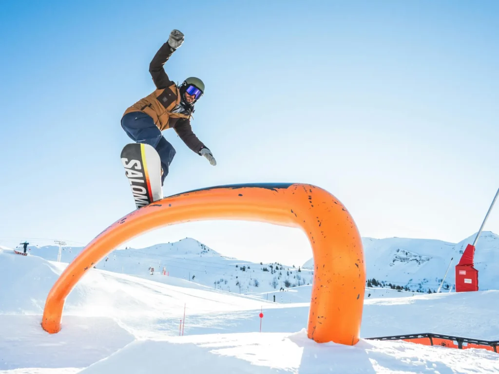 Ein Snowboarder vollführt einen Sprung über ein orangefarbenes Rail, ein hoher Sprung mit erhobener Hand, im Hintergrund winterliche Berge.